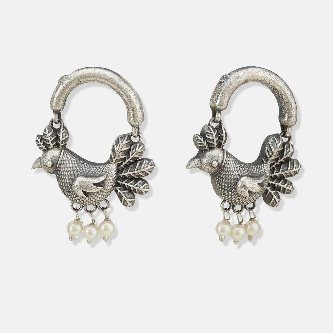 A pair of silver-toned huggie hoop earrings featuring a detailed bird charm with leafy accents and small dangling pearl beads, shown on a light gray background.