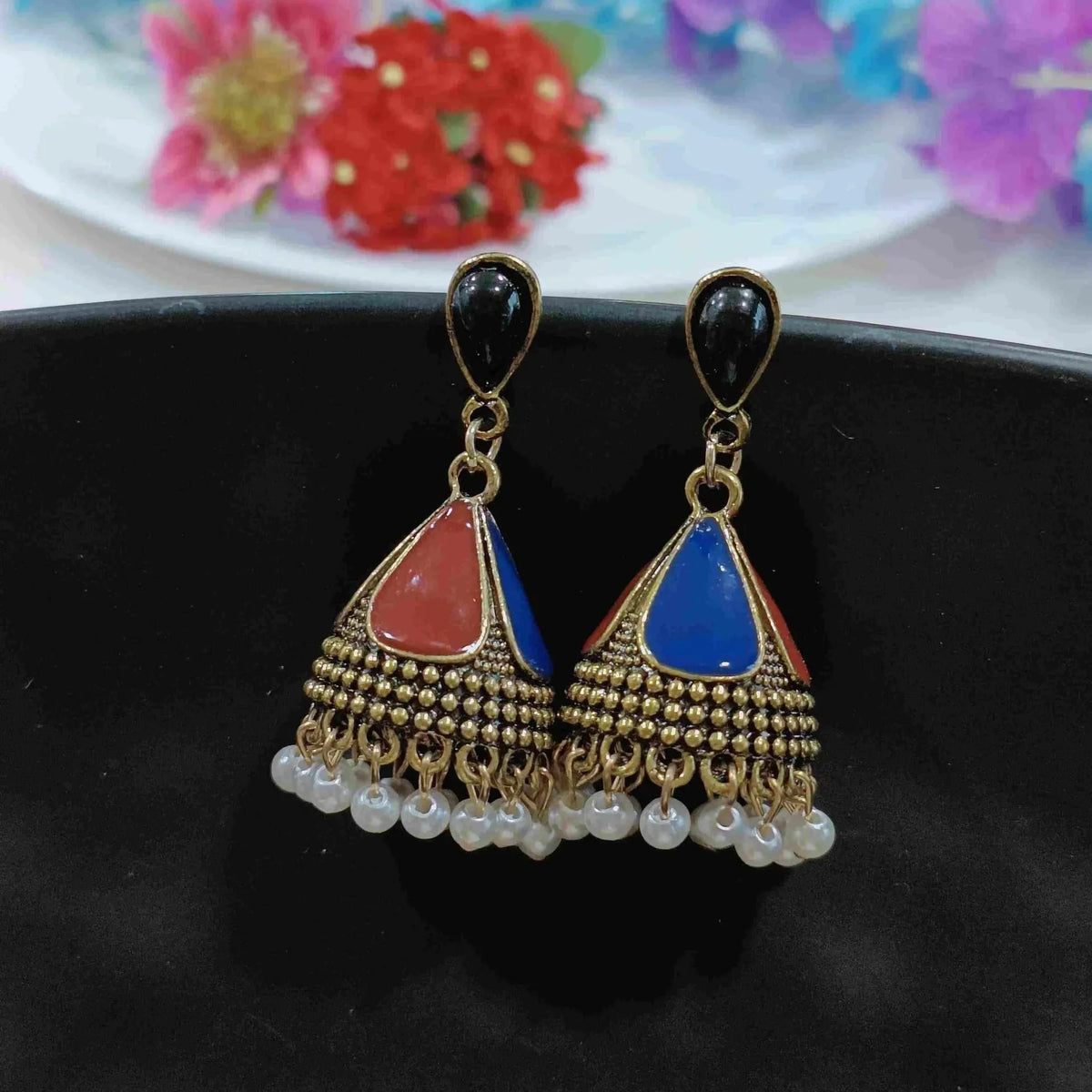 A pair of ornate gold-toned jhumka earrings with black teardrop-shaped studs, red and blue enamel details, and dangling white pearl beads.