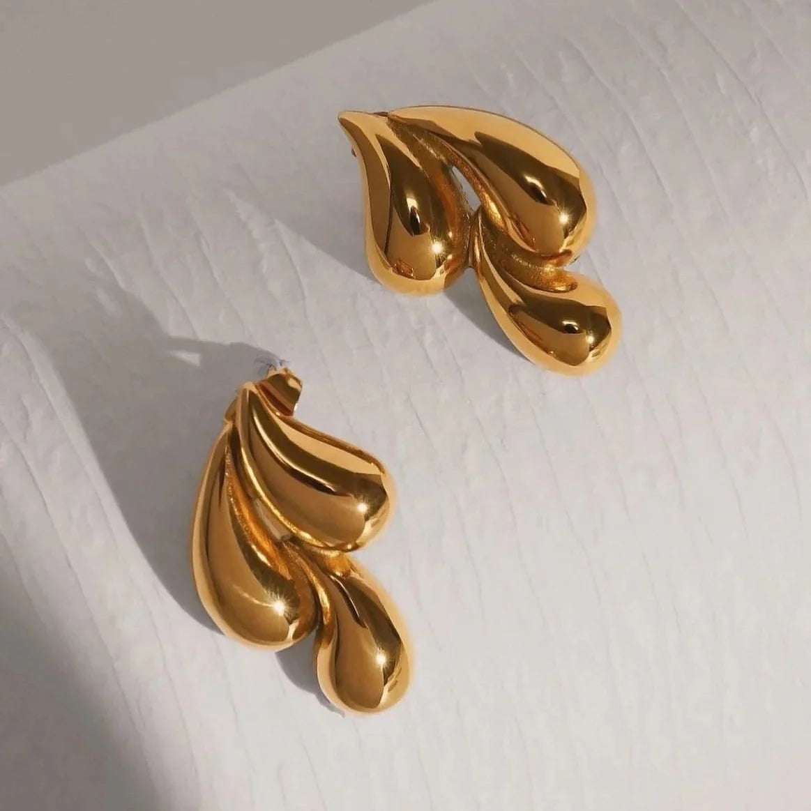 A pair of shiny gold teardrop-shaped earrings with a glossy, reflective finish, displayed on a textured white surface.