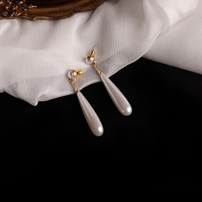 A pair of gold-tone drop earrings with teardrop-shaped white faux pearls, styled on draped white fabric over a black background.