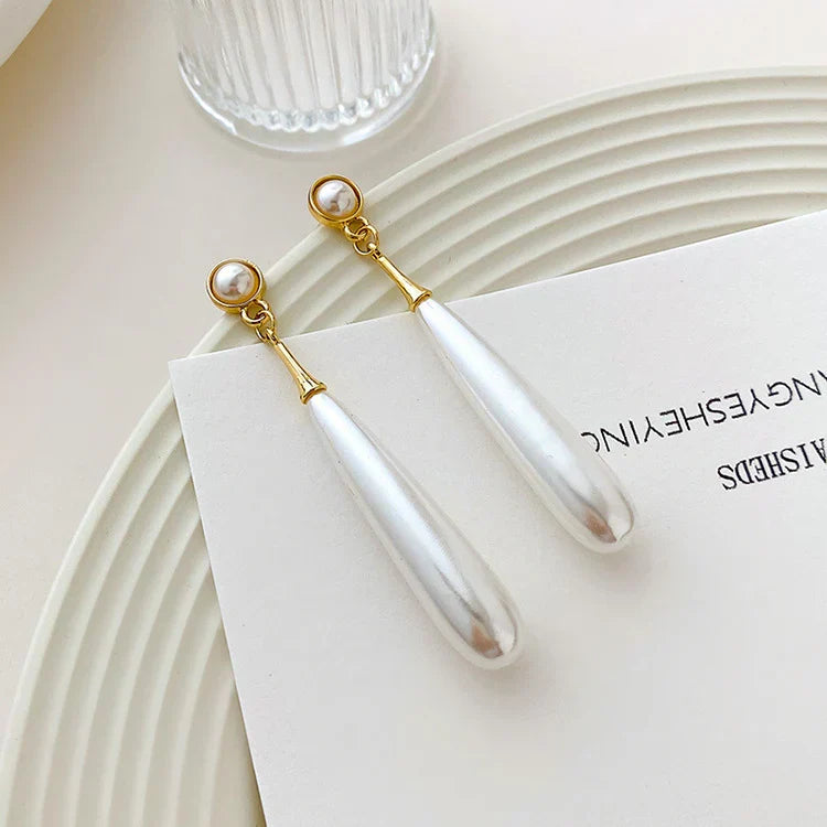 A pair of long teardrop-shaped drop earrings with white pearl-like drops and gold-toned metal connectors, resting on a white card over a white ridged plate.