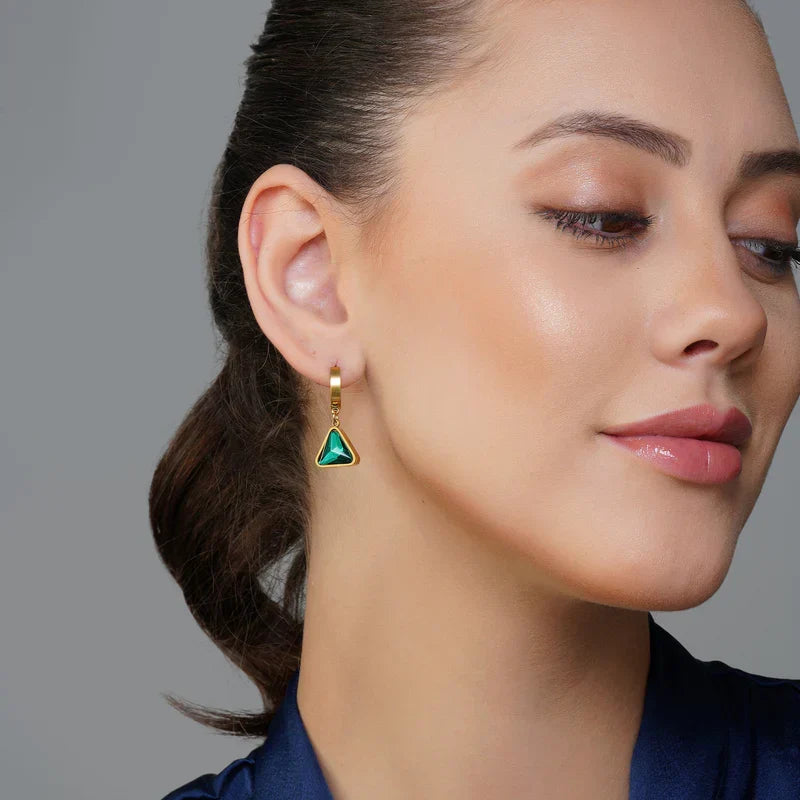 A close-up of a woman wearing a gold hoop earring with a triangular green stone, shown against a neutral gray background.