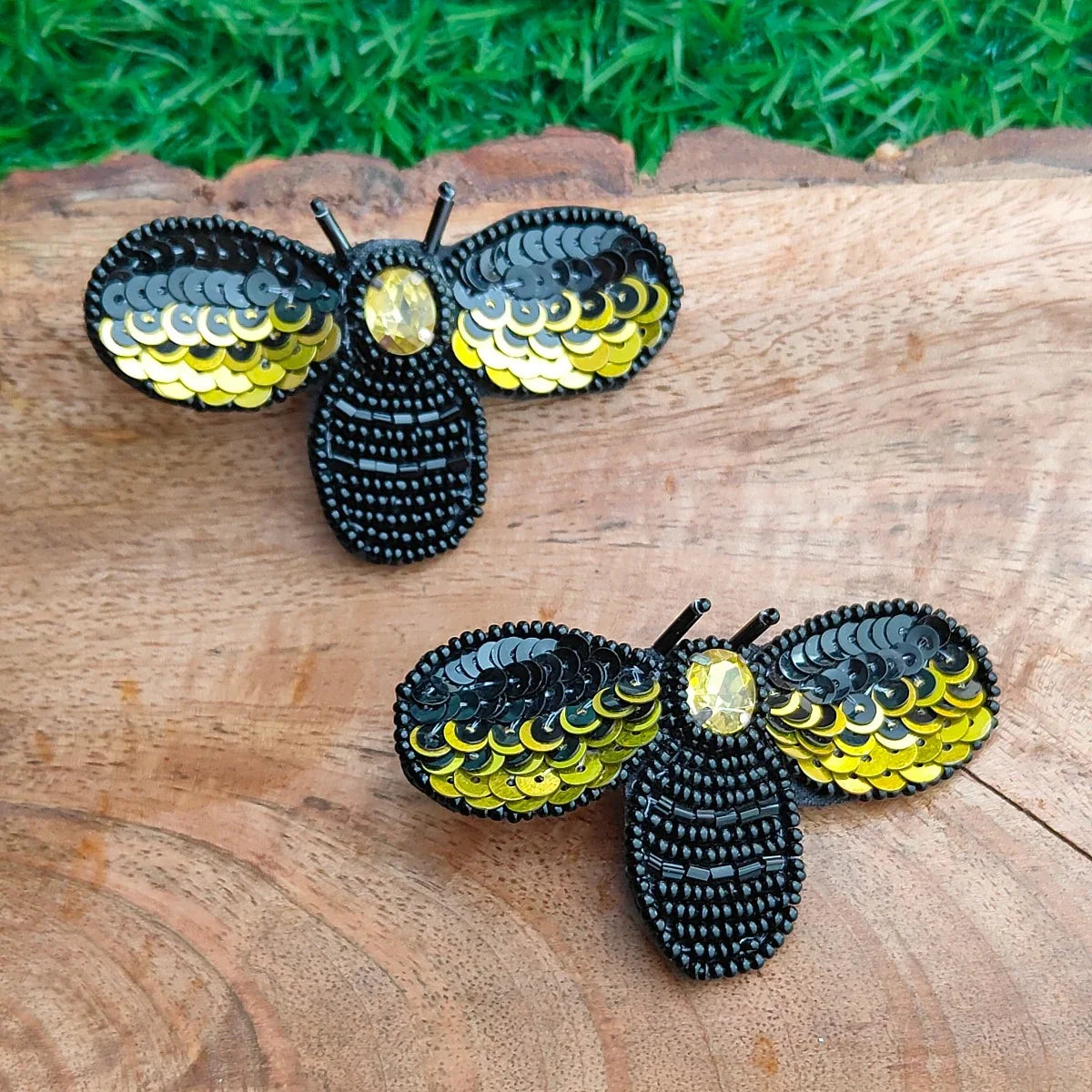 Two black beaded bumblebee earrings with yellow sequined wings, displayed on a rustic wooden surface with green grass in the background.