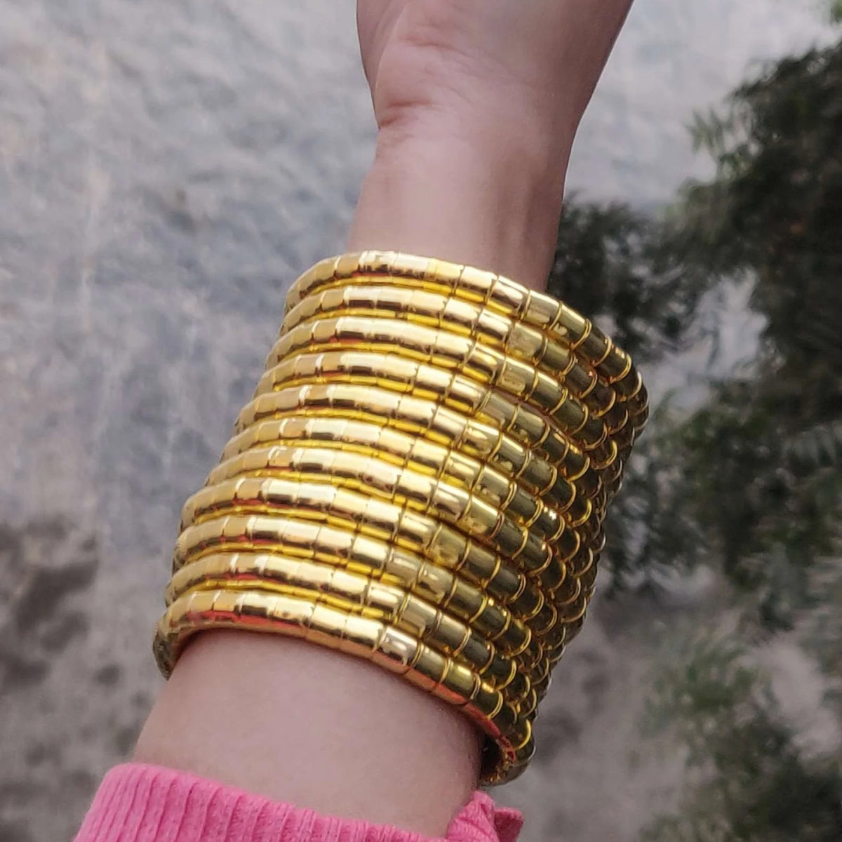 A close-up of a person's wrist wearing a stack of wide, chunky gold bangles with a textured, segmented design. The person is wearing a pink knit sweater, and the background shows a blurred outdoor scene with gray stone and green foliage.