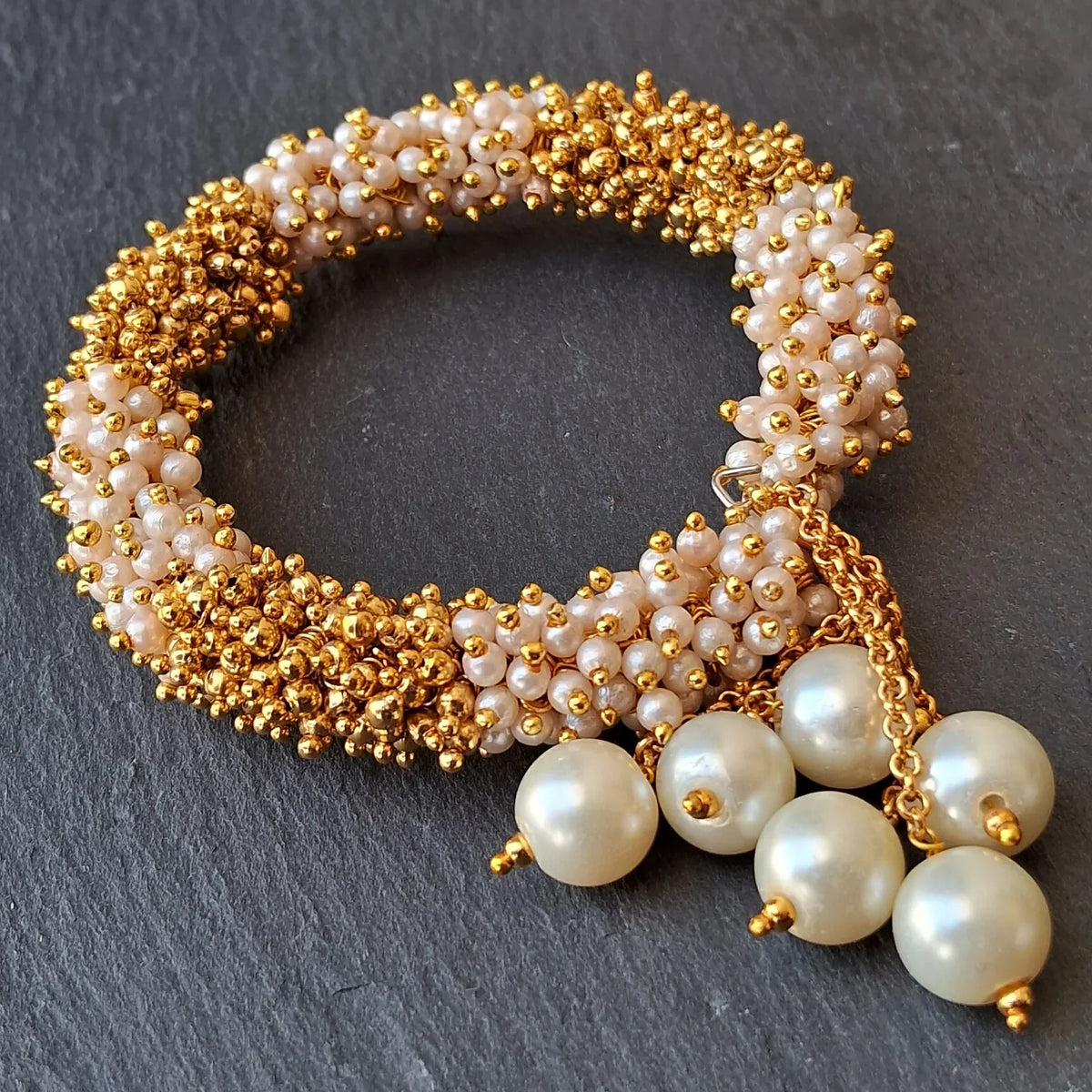 A beaded bracelet with gold-tone seed beads, pale pink pearl-like beads, and three dangling cream pearl charms on a dark gray slate background.