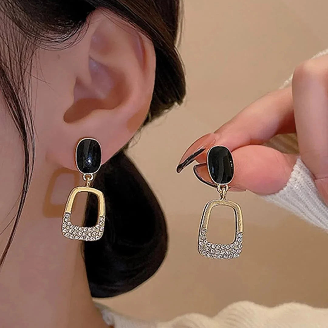 A close-up lifestyle shot of a model wearing and holding a pair of dangling earrings. The earrings feature a polished gold-tone geometric frame with a row of clear rhinestones and a black oval stone at the top.