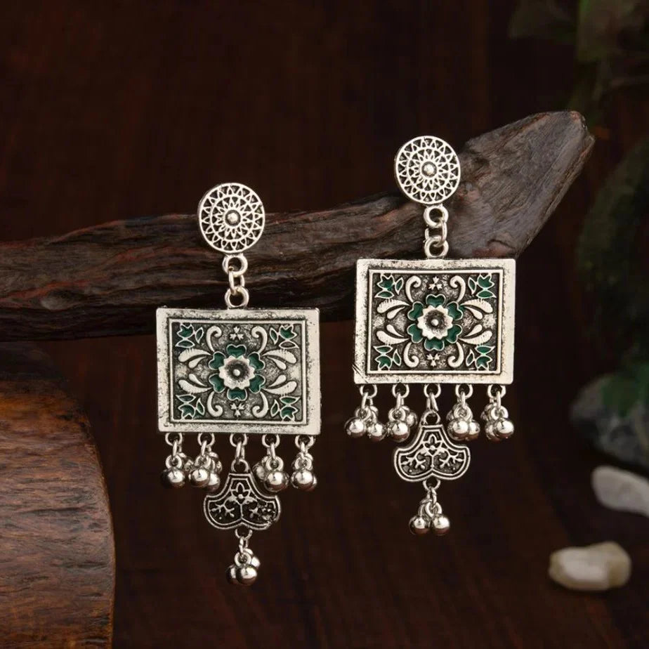 A pair of ornate silver-toned dangle earrings with square green enamel floral panels and dangling silver bells.