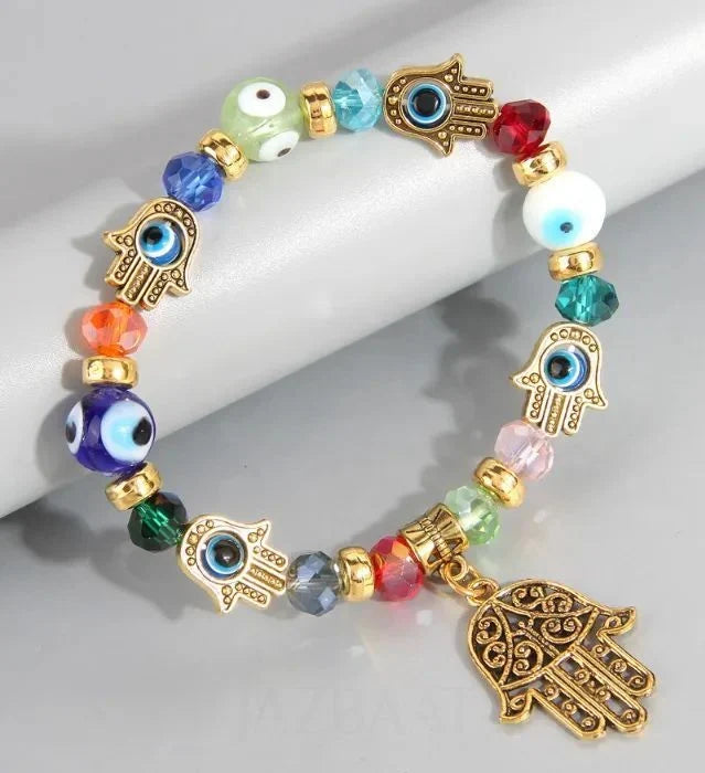A beaded stretch bracelet with gold-tone Hamsa charms, evil eye beads, and faceted crystal beads in multiple colors, displayed on a light gray cylindrical stand.