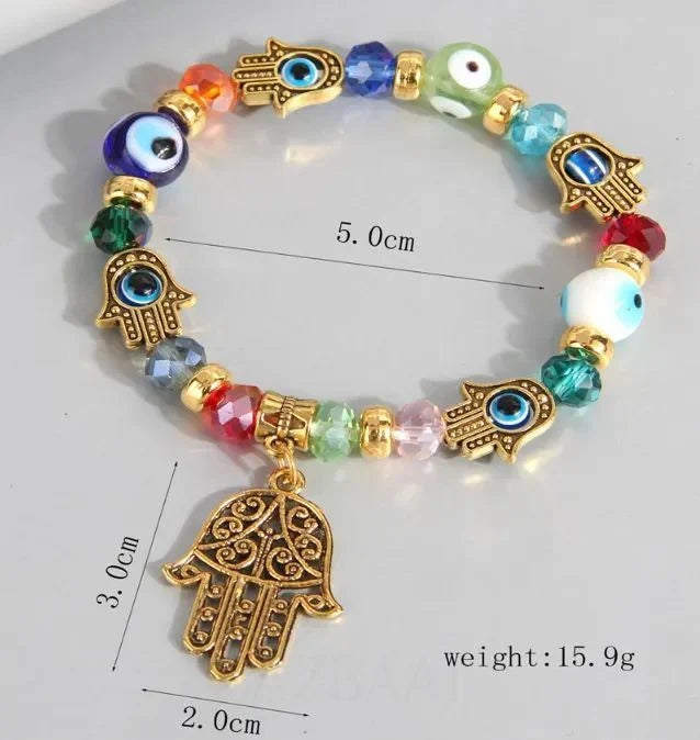 A gold-tone Hamsa hand charm bracelet with evil eye beads and gold spacer beads, shown on a light gray background with measurement annotations indicating 5.0cm width, 3.